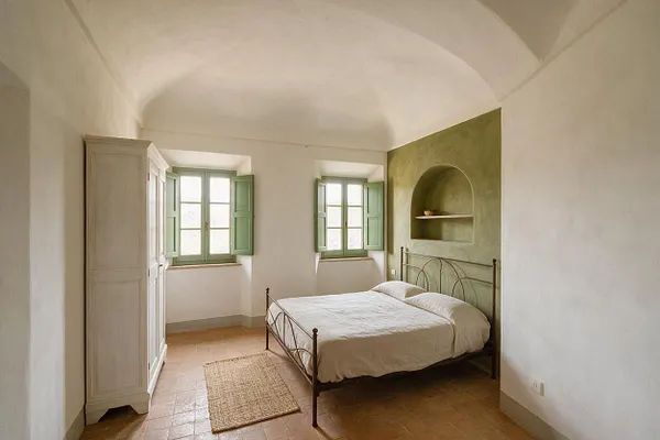 A serene Mediterranean-style master bedroom with a white vaulted ceiling, terracotta floor tiles, and white plaster walls. The room features a wrought-iron bed, a pale wood wardrobe, two mint-green shuttered windows, and a green accent wall with an arched niche shelf.