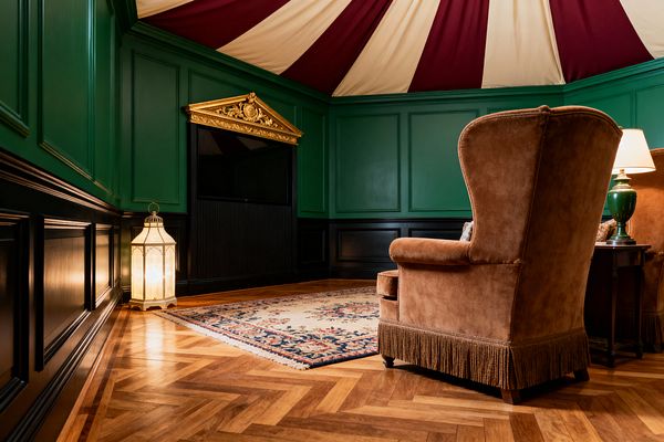 Traditional media room with emerald green paneled walls and black wainscoting centers on a built-in television crowned by a gilded neoclassical pediment beneath a burgundy-and-ivory tented ceiling. A caramel velvet wingback with bullion fringe faces a cream, floral-bordered wool rug on herringbone oak parquet, while a glass-and-brass floor lantern and a green ceramic table lamp cast warm, intimate light.