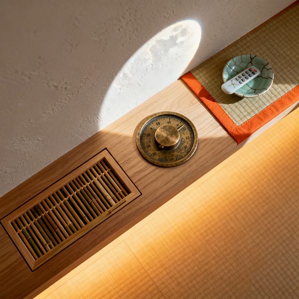Close-up of a Japanese Zen–style media room detail. A light oak ledge abuts a warm white textured plaster wall. Set into the wood are a narrow bamboo slatted grille and a round patinated brass dial with numeric markings. Beside them, a tatami mat with bright orange fabric edging holds a small celadon dish containing a white remote. Soft sunlight creates a circular glow across the surface, emphasizing natural textures and calm simplicity.
