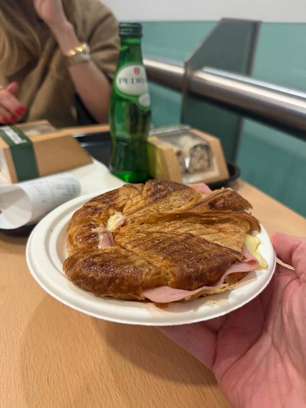 A hand holds a warm ham and cheese croissant on a plate, with a bottle of sparkling water and a woman in the background.