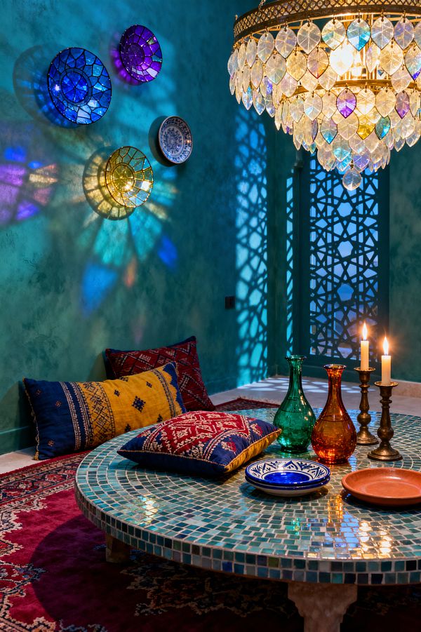 A jewel-toned Bohemian dining nook with a low, round table tiled in teal mosaic set on a crimson Persian rug, surrounded by patterned kilim floor cushions. Teal plaster walls, a Moorish lattice screen, and a brass chandelier with opalescent drops create colorful shadows, while the table holds blue-and-white ceramics, a terracotta dish, green and amber glass bottles, and brass candlesticks with lit candles.