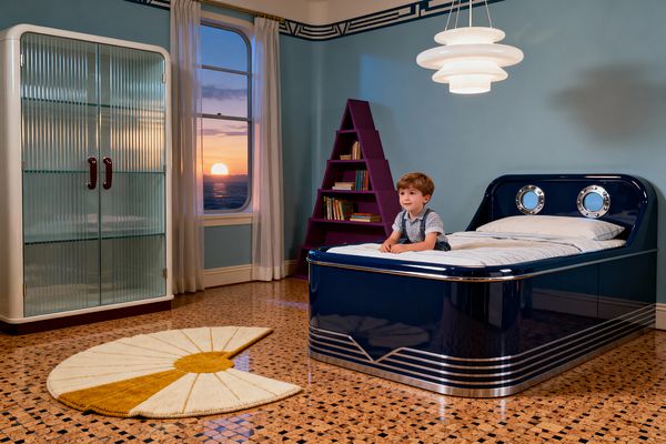 A kids' bedroom in Art Deco style features a glossy navy bed with chrome banding and two round porthole details in the headboard, where a child sits. To the left is a cabinet with rounded corners and fluted glass doors with burgundy pulls; at the back a triangular purple bookcase stands against pale blue walls with a navy geometric frieze, beneath an arched window with sheer curtains and a sunset view; a stepped white pendant hangs over terrazzo flooring and a cream-and-mustard fan-shaped rug.