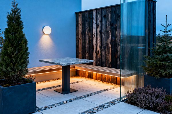 A compact Scandinavian-style patio features light gray concrete pavers separated by river pebbles, a square granite-topped table on a black pedestal, and a built-in slatted wood bench. Warm LED lighting under the bench glows against a charred timber privacy wall and a blue stucco facade with a round sconce, while a vertical fluted glass screen and dark planters with evergreens and heather add texture and privacy.