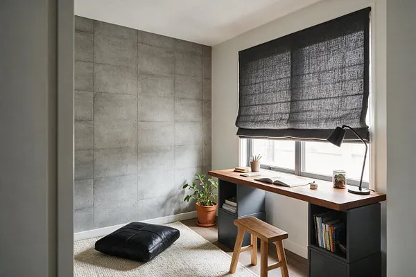 A compact sunroom or study nook features a raw concrete accent wall, a long wood-topped desk with dark storage cubbies, and a black desk lamp under a large window. The space includes a woven cream rug, a black floor cushion, a small wooden stool, a potted plant, and a charcoal Roman shade filtering daylight.