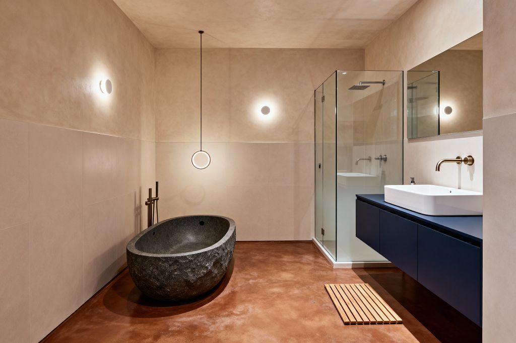 A minimalist modern bathroom features a rough-hewn oval stone soaking tub beside a freestanding brushed-brass filler, a slim ring pendant, and two small wall sconces. Opposite, a frameless glass shower and a floating matte-navy vanity with a white vessel sink sit against warm beige plaster and large-format tile walls, above a terracotta-toned polished concrete floor with a slatted wood bath mat and a wide mirror reflecting the soft, warm light.