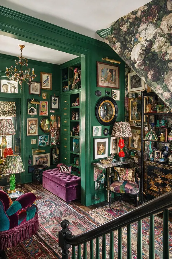 A maximalist entryway with deep emerald green walls, built-in shelving, and a staircase wrapped in large-scale floral wallpaper. The space is filled with framed artwork, mirrors, patterned rugs, velvet seating, colorful lamps, and decorative objects layered throughout.