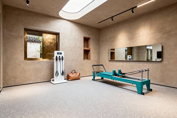A minimalist home gym with warm sand-colored plaster walls features an elongated oval skylight and a brass-framed window that bring in soft natural light. A teal Pilates reformer sits on light gray speckled rubber flooring opposite a long, rounded-edge wall mirror, with black track lighting above, a white equipment tower with resistance cords, two recessed oak niches, and a tan leather duffel completing the calm, contemporary space.