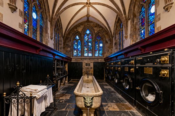 A dramatic laundry room set in a Gothic-style chamber with ribbed vaulted ceilings, pointed stone arches, and tall stained-glass lancet windows glowing red and blue. A carved stone trough with a gargoyle spout sits center, flanked by black paneled wainscoting. Industrial black washers with brass hardware line the right wall, with vintage iron piping and a towel rack on the left. The floor is dark slate and the lighting is moody and warm.