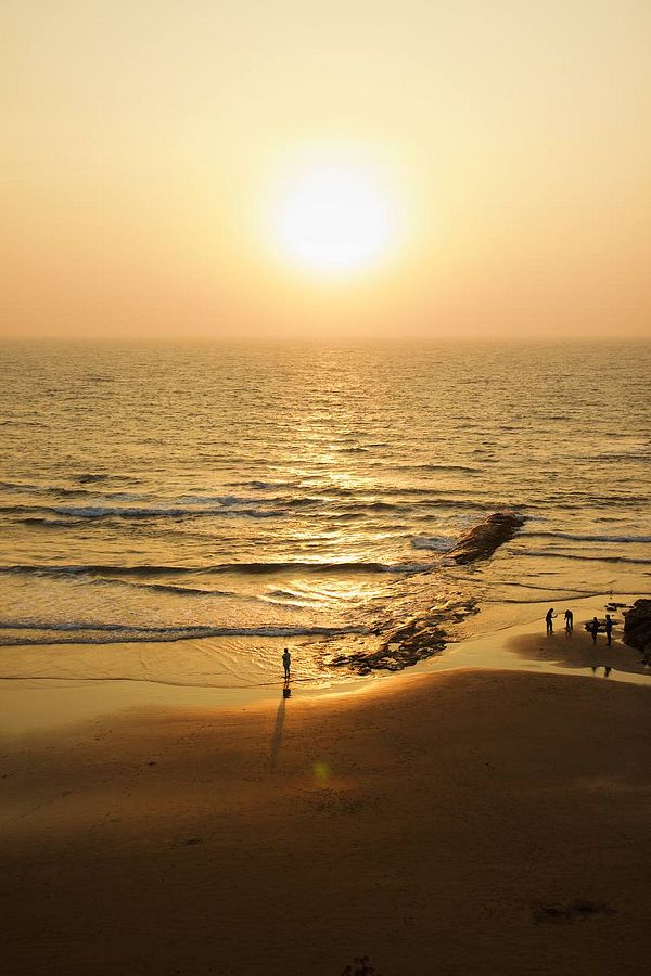A serene beach scene at sunset in Goa, featuring a lone figure and small groups of people along the shore.