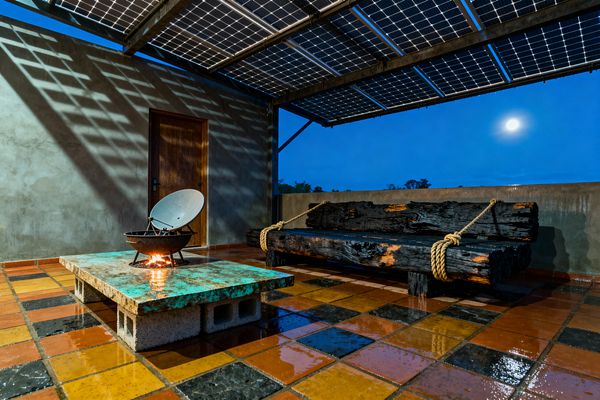 Nighttime patio with a metal-framed canopy of solar panels casting a grid of shadows over wet checkerboard tiles in terracotta, ochre, and charcoal. A low table made from a green-patinated metal slab on concrete blocks holds a steel fire bowl with a repurposed dish, while a bench built from charred timber logs, tied with thick rope, sits against smooth gray plaster walls and a wooden door, with the moon visible beyond.