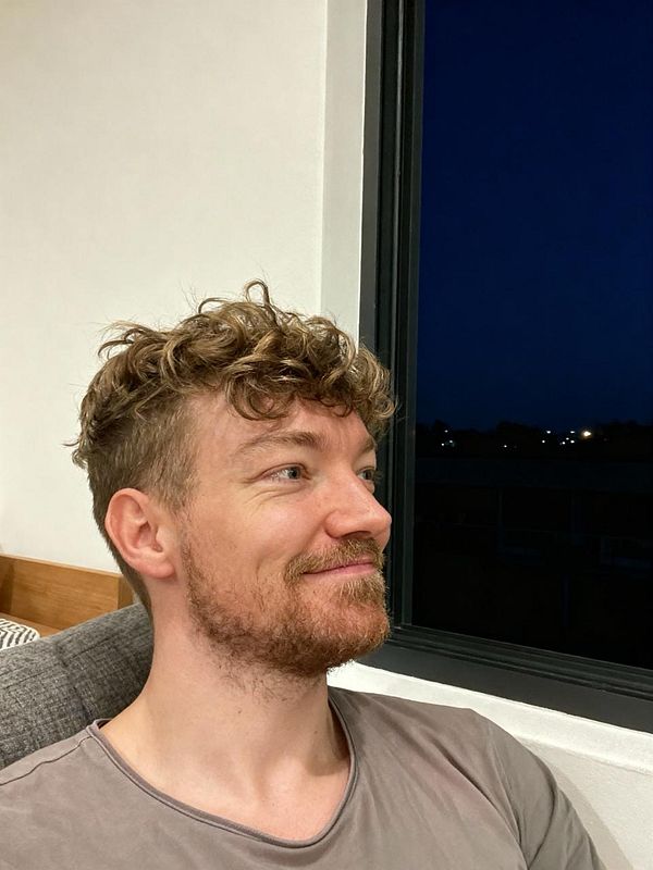 A man with curly hair and a beard is sitting by a window, smiling softly.