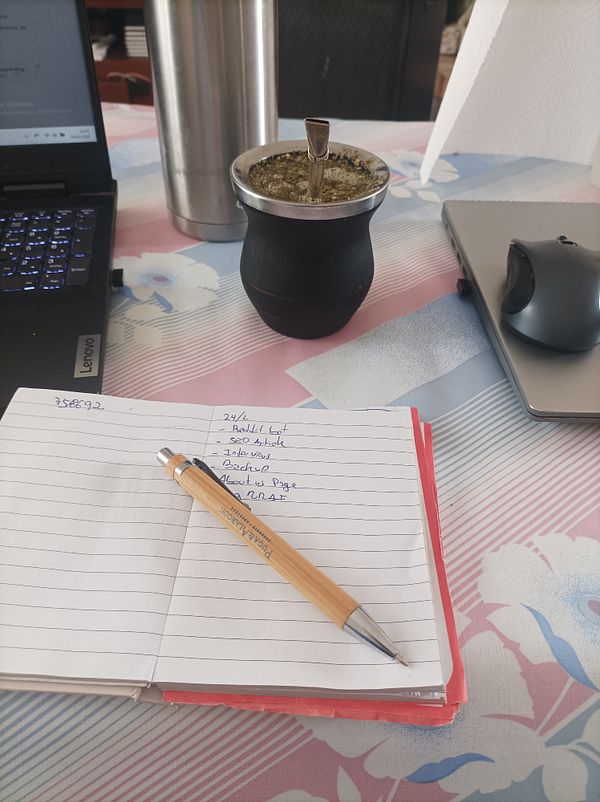 A workspace featuring a laptop, a notebook with a to-do list, a mate cup, and a thermos.