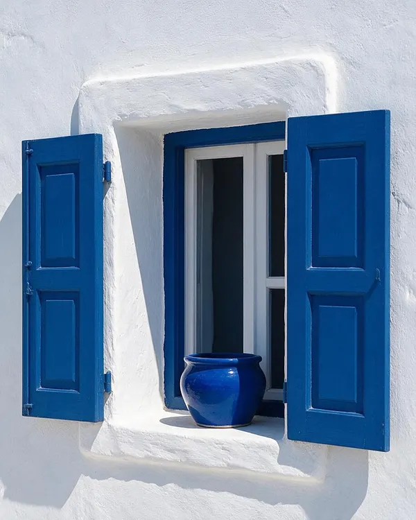 A white stucco Mediterranean exterior wall with a small recessed window framed in bright cobalt blue. The window has matching blue wooden shutters opened outward, and a glossy blue ceramic pot sits on the sill, creating a simple, sunlit contrast.