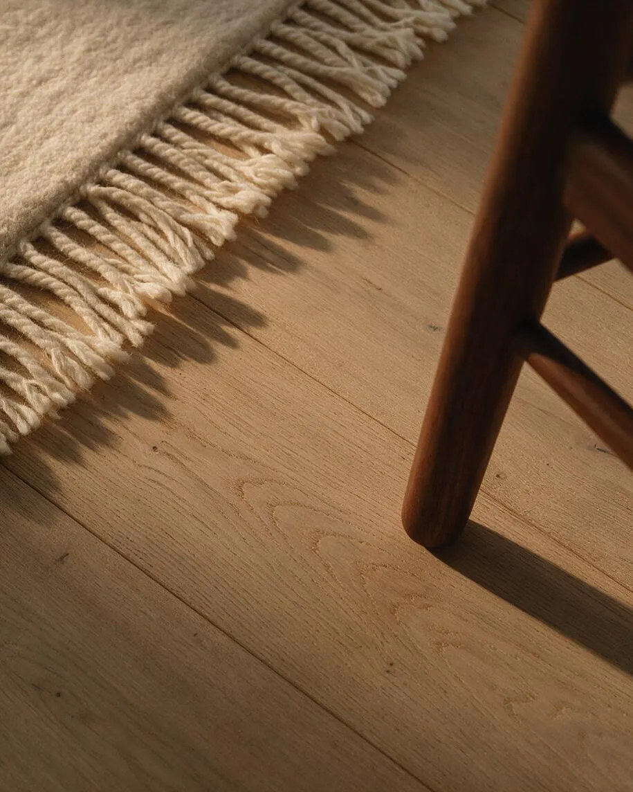 A close-up view of a cream wool rug with thick fringe resting on light oak floorboards. A dark wooden chair leg enters from the right, and warm sunlight casts soft shadows across the natural wood grain.