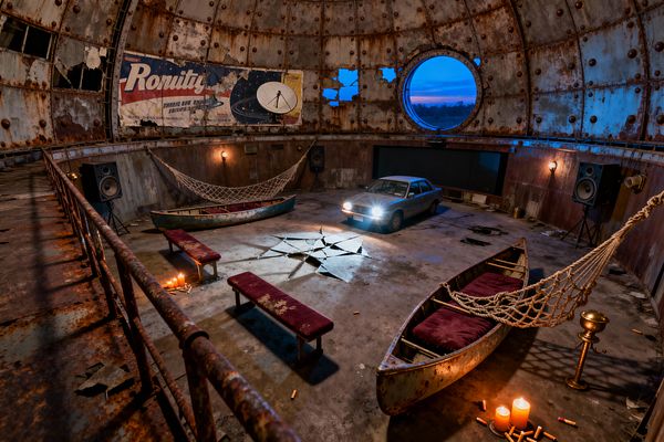 Inside a rusted, dome-shaped chamber, a silver sedan with headlights on faces a wall-mounted screen between large speakers. The concrete floor features a star-shaped crack, while metal canoes with burgundy cushions serve as seating, linked by thick rope hammocks; distressed benches, burning candles, a brass pedestal, a faded space-age billboard, and a round porthole window glowing blue at dusk complete the improvised media room.