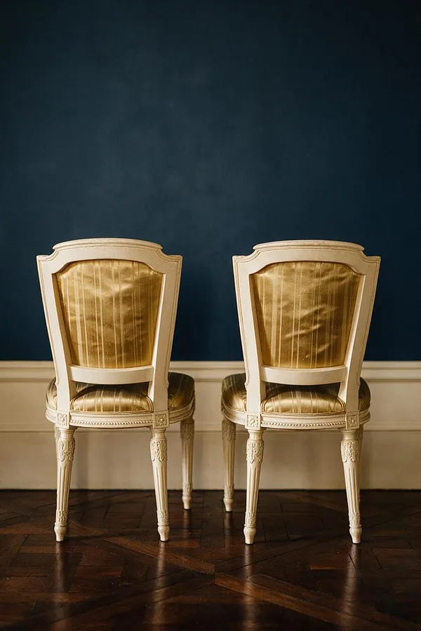 Two traditional upholstered side chairs are positioned side by side against a deep blue wall. Each chair has a painted cream wood frame, a curved crest rail, gold silk-like upholstery, carved legs, and a dark herringbone wood floor below.