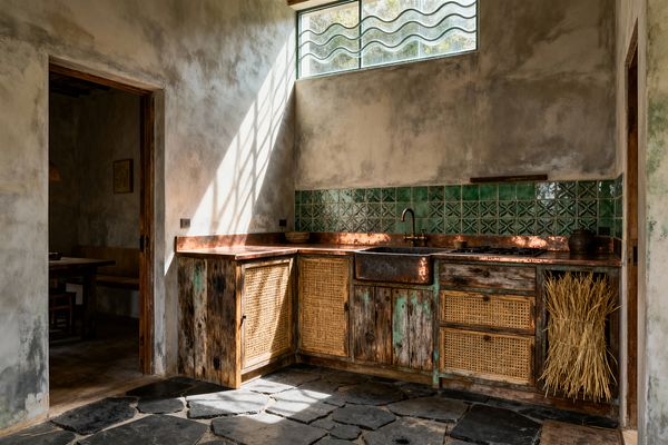 Rustic kitchen with L-shaped base cabinets made from weathered reclaimed wood and woven cane panels. A patinated copper farmhouse sink and matching countertop sit beneath a high clerestory window with wavy glass, casting diagonal light. The backsplash is hand-glazed green tiles, some with embossed motifs. Walls are mottled limewash plaster and the floor is irregular dark slate. An aged brass faucet, ceramic vessels, and a bundle of wheat complete the timeworn look.
