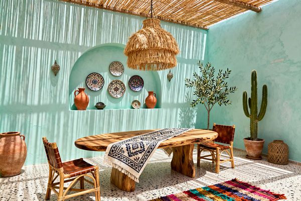 A bright bohemian dining room with mint plaster walls and a slatted reed roof casting linear shadows. A live-edge oval wood table with a navy-and-ivory runner is paired with two rattan chairs with leather seats; an arched wall niche displays patterned ceramic plates and terracotta jugs. A raffia pendant hangs above, terrazzo flooring grounds a multicolor rug, and potted cactus and an olive tree add greenery.
