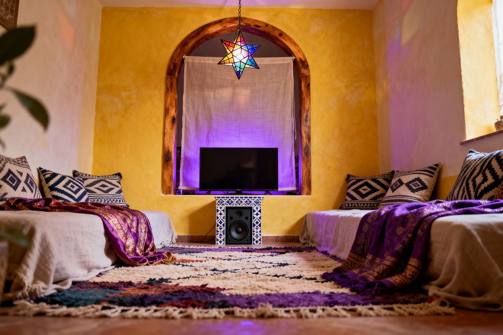 A bohemian media room with two low sofas facing each other, covered in natural linen and layered with indigo-and-ivory patterned cushions and purple, gold-trimmed throws. At the center, an arched niche with rustic wood trim holds a TV on a blue-and-white tiled stand; a linen curtain and violet backlighting sit behind it. A cream, multicolor shag rug spans terracotta floors, while a multicolored stained-glass star pendant casts jewel-toned reflections across sunny yellow plaster walls and a small side window.