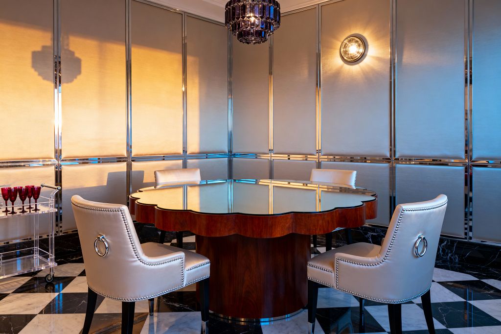 A vintage glam dining corner features a scalloped walnut pedestal table with a mirrored top, surrounded by four cream leather chairs detailed with silver nailheads and ring pulls. The floor is black-and-white marble in a large checkerboard. Champagne-colored wall panels with polished chrome trim reflect a smoky crystal drum chandelier and a round prismatic sconce. At left sits a clear acrylic bar cart with ruby glasses, while warm light casts soft reflections across the room.