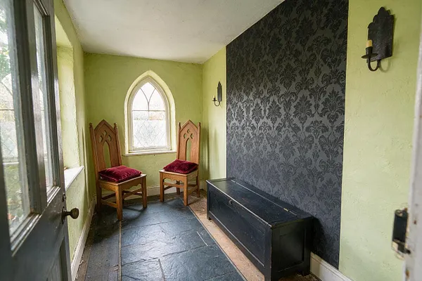 A small Gothic-style front porch with pale green walls, a pointed arched window at the back, and two carved wooden chairs with burgundy cushions. One wall is covered in dark damask wallpaper, and a long black storage bench sits below it on a slate-tiled floor.