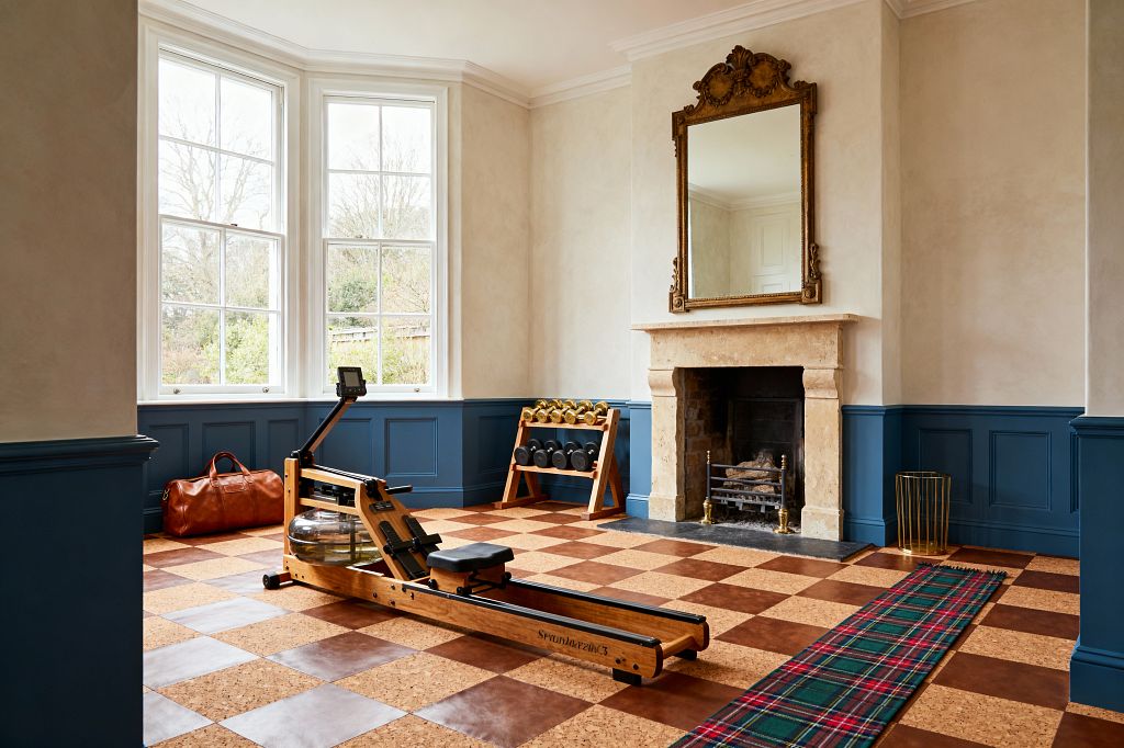 A light-filled home gym in a traditional room with deep blue paneled wainscoting, ivory plaster walls, and a carved limestone fireplace topped by an ornate gilded mirror. A wooden water rowing machine rests on a checkerboard floor of cork and brown tiles beside a rack of black dumbbells and brass kettlebells, a red-green tartan runner, a tan leather duffel, and tall white sash windows with garden views.