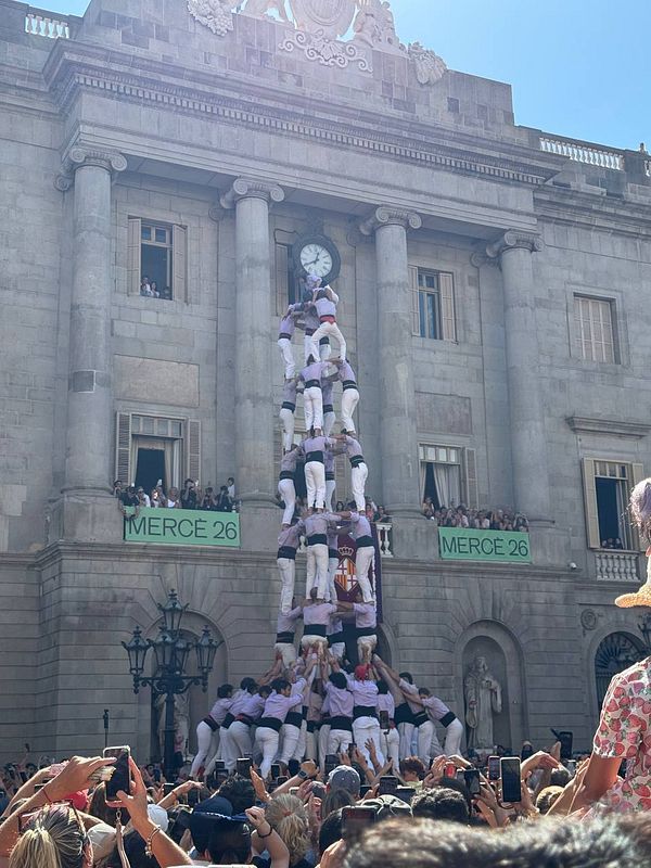 A tall human tower is formed in front of a historic building with a large crowd watching and capturing the moment.