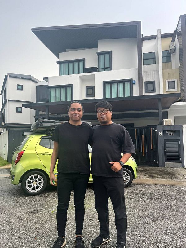 Two individuals pose in front of a modern house with a green car parked nearby.