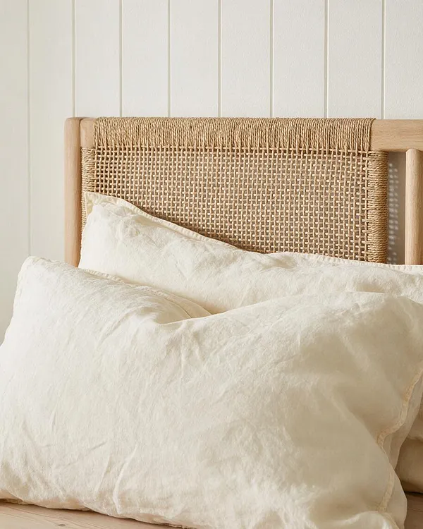 A coastal-style bed features a light wood frame and a woven cane headboard panel behind layered cream-colored linen pillows. The background is a simple white paneled wall, and the overall look is airy, natural, and relaxed.