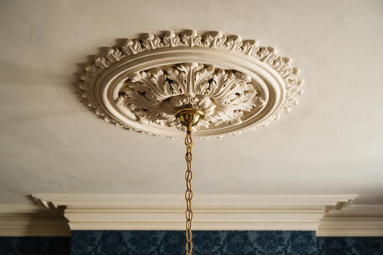 An ornate Victorian-style ceiling detail with a carved plaster medallion featuring layered oval moldings and leaf motifs. A brass ceiling rose at the center supports a hanging chain, with cream crown molding and deep blue patterned wallpaper below.