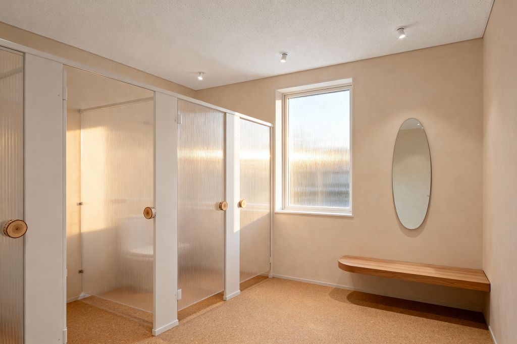 A minimalist home gym changing area includes three private cubicles with translucent fluted panels set in white frames, each fitted with round natural-wood pulls. Warm beige plaster walls and cork flooring are lit by a window of reeded glass that diffuses daylight, while small ceiling spotlights add even brightness. Along the opposite wall, a floating solid-wood bench sits beneath a tall frameless oval mirror, with the overall palette kept neutral and uncluttered.