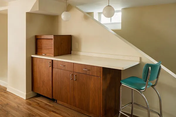 A mid-century modern basement nook features a built-in walnut bar cabinet with a light countertop beneath a sloped stairway. A teal vinyl barstool with chrome tubing sits beside warm beige walls, wood-look flooring, and two globe pendant lights overhead.