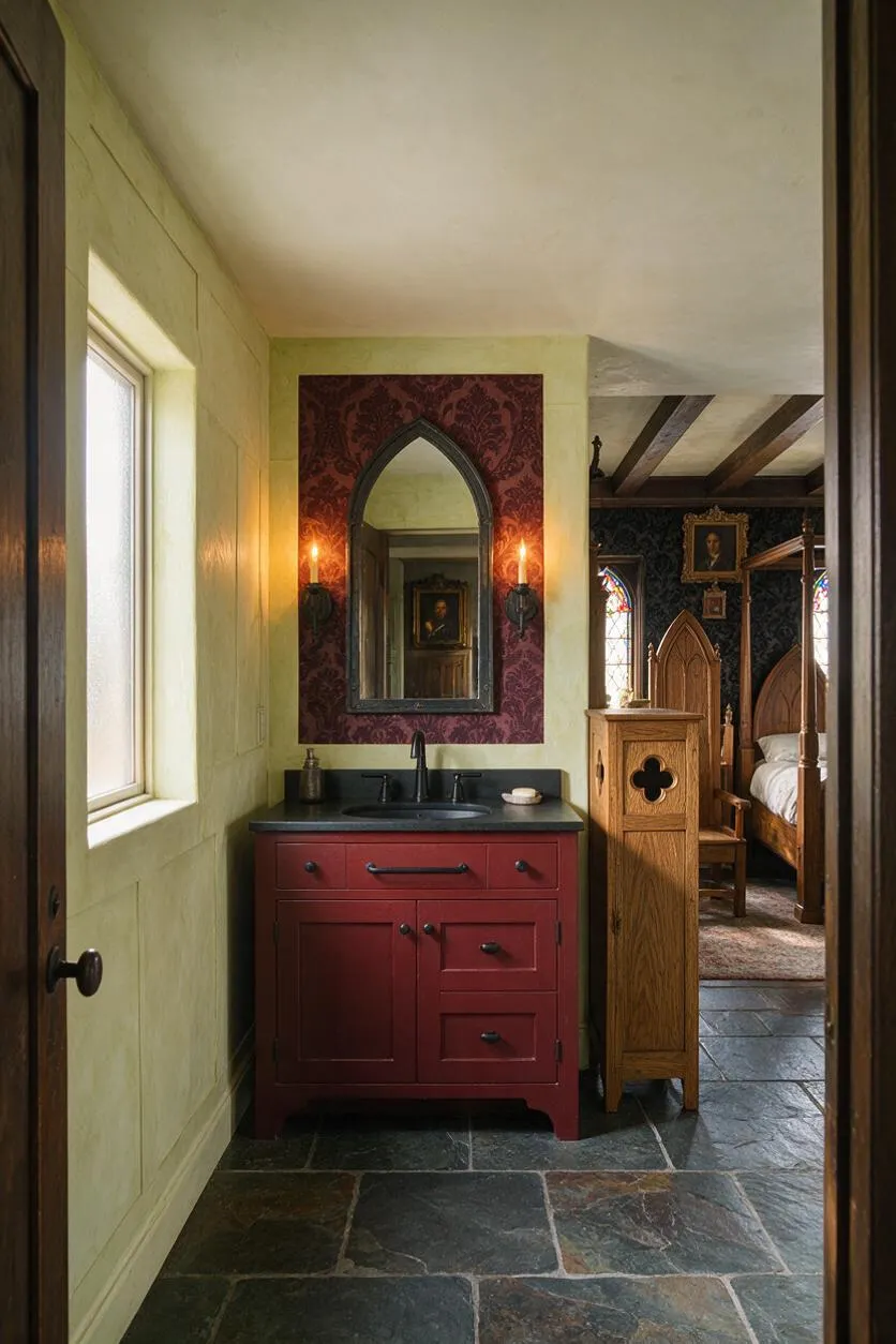A Gothic-style master bathroom with a deep red vanity, black countertop, and arched mirror framed by patterned burgundy wallpaper. Stone tile flooring, candle-style sconces, and a glimpse into a matching bedroom with carved wood furniture and stained-glass details complete the moody, historic atmosphere.