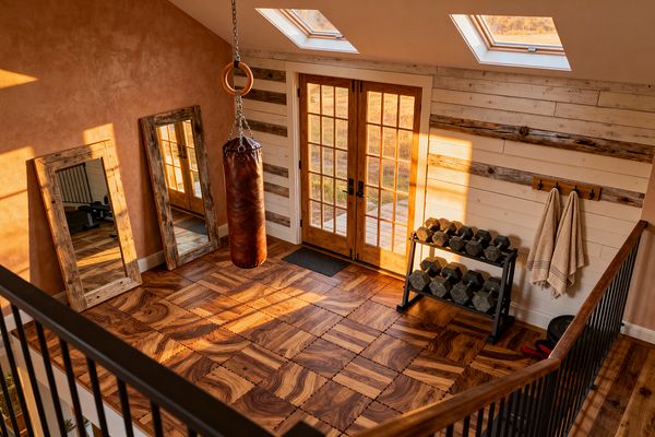 A farmhouse-style home gym bathed in natural light with interlocking wood‑grain floor tiles and a brown leather punching bag hanging from a chain beneath two skylights. Two large mirrors with reclaimed wood frames lean on a warm clay‑plaster wall, while the opposite wall features white shiplap with weathered wood bands, natural-wood French doors, a black metal dumbbell rack of hex weights, and a wooden peg rail with beige towels.