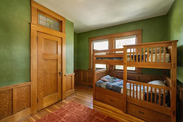 A Craftsman-style kids' room with green walls, warm wood wainscoting, and a built-in wooden bunk bed along the right side. A paneled wood door with a small stained-glass transom sits on the left, while windows above the bunk bed bring in natural light.