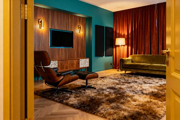 View through a mustard door into a mid-century modern media room with teal walls, a walnut slat TV wall, and brass sconces. A walnut-and-terrazzo credenza sits beneath the screen beside an Eames-style black leather lounge and ottoman; opposite are an olive green velvet sofa, rust velvet floor-to-ceiling drapes, black acoustic panels, a brass floor lamp with a drum shade, oak herringbone flooring, and a thick brown-and-cream shag rug.