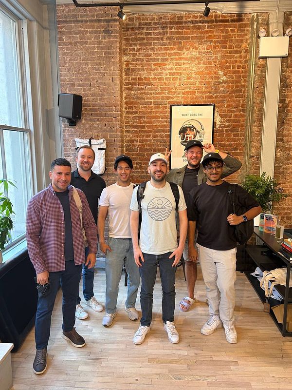 A group of six individuals poses together in a stylish office space with a brick wall backdrop.