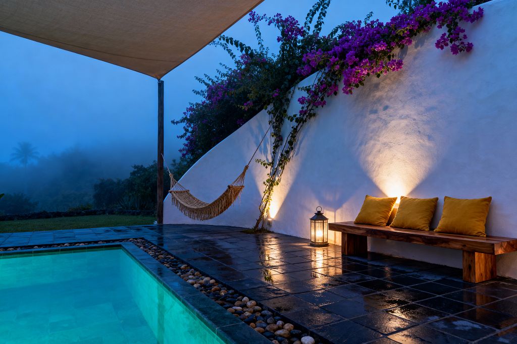 A tropical patio at dusk features a glowing turquoise pool beside glossy dark stone pavers with a pebble border. A curved white stucco wall is washed by warm uplights and topped with trailing magenta bougainvillea. A solid wood bench with three mustard cushions sits next to a lit metal lantern, while a beige shade sail shelters a knotted macramé hammock. Misty palms and dense greenery loom beyond, creating a tranquil, resort-like outdoor retreat.