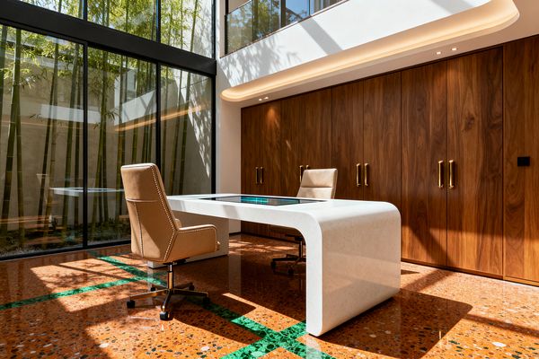 A double-height home office features a curved white solid-surface desk with a central glass inset and two tan leather swivel chairs. One wall is clad in floor-to-ceiling walnut cabinetry with brass handles, while the opposite side is full-height black-framed glass overlooking a bamboo courtyard; terra-cotta terrazzo flooring with green marble inlay and warm LED cove lighting complete the clean, futuristic space.