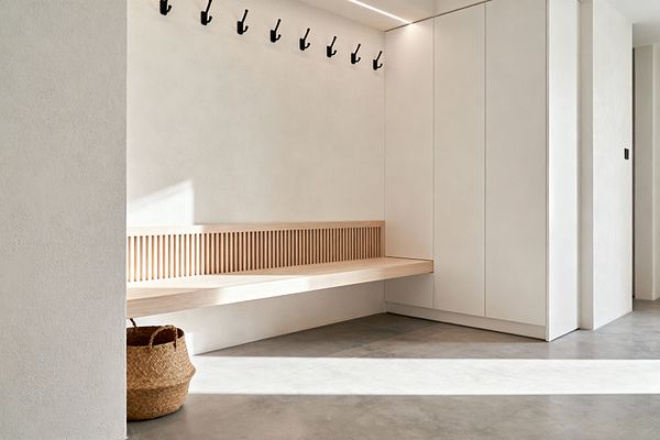 A minimalist mudroom shows a floating light-oak bench with a vertical slatted back along a warm white wall beneath a line of matte black coat hooks. At the right, full-height, handleless white cabinets meet the bench, while a woven basket sits under the seat. The floor is light gray concrete with daylight streaming from the left, and a slim recessed ceiling light adds soft illumination. The space is tidy, neutral, and highly functional.