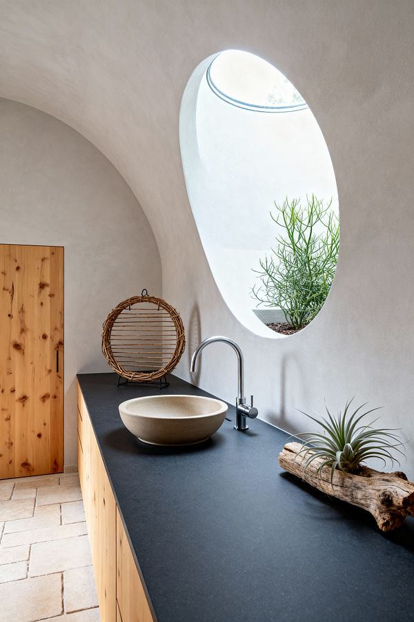 A biophilic laundry workspace with a long matte-black countertop, stone vessel sink, and chrome faucet sits under a sweeping curved plaster wall and ceiling. An oversized oval window floods the space with daylight and frames green foliage outside, while honey-toned knotty pine cabinets, a wicker accent, an air plant on driftwood, and pale limestone floor tiles create a calm, organic atmosphere.