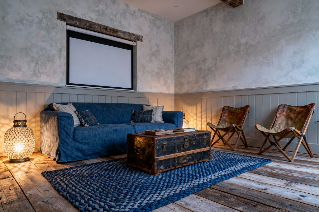 A cozy farmhouse-style media room with a deep indigo slipcovered sofa facing an antique steamer trunk used as a coffee table on a braided blue rug. A retractable projector screen sits above gray-green tongue-and-groove wainscoting and mottled plaster walls, with a rustic timber beam above. Two folding cognac leather chairs line the right wall, while glass-and-metal lanterns on the floor cast a warm patterned glow across weathered plank wood flooring.