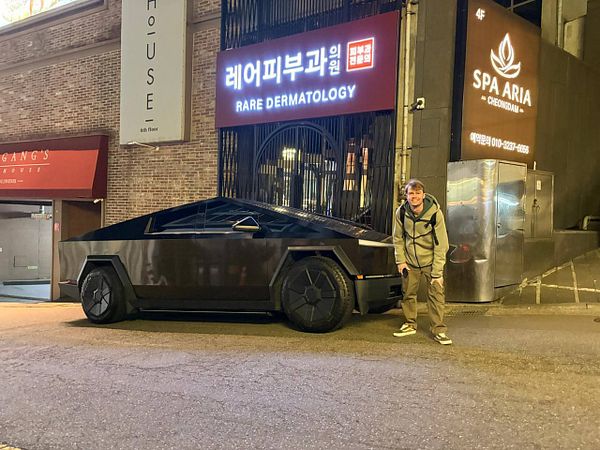 Silver Tesla Cybertruck parked outdoors near dark cars under trees with a clear sky in the background.