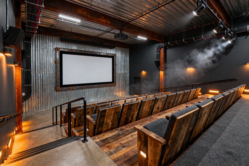 A stepped media room features rows of reclaimed wood theater seats with gray upholstered cushions facing a large projection screen framed in riveted steel on corrugated metal cladding. The space has exposed beams, a ribbed metal ceiling, and visible conduit with track lights, plus a ceiling projector. Concrete floors, charcoal-painted walls, wall speakers, and amber step lights create a moody glow, with a light haze accentuating the directional spotlights.