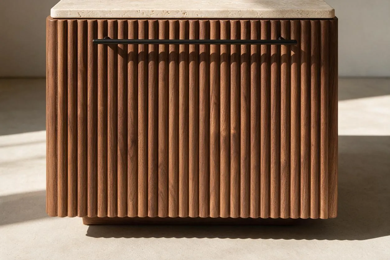 A modern cabinet with vertical fluted wood paneling in a warm brown finish and a light stone top. A slim black handle spans the upper front, and soft sunlight casts shadows across the textured surface.