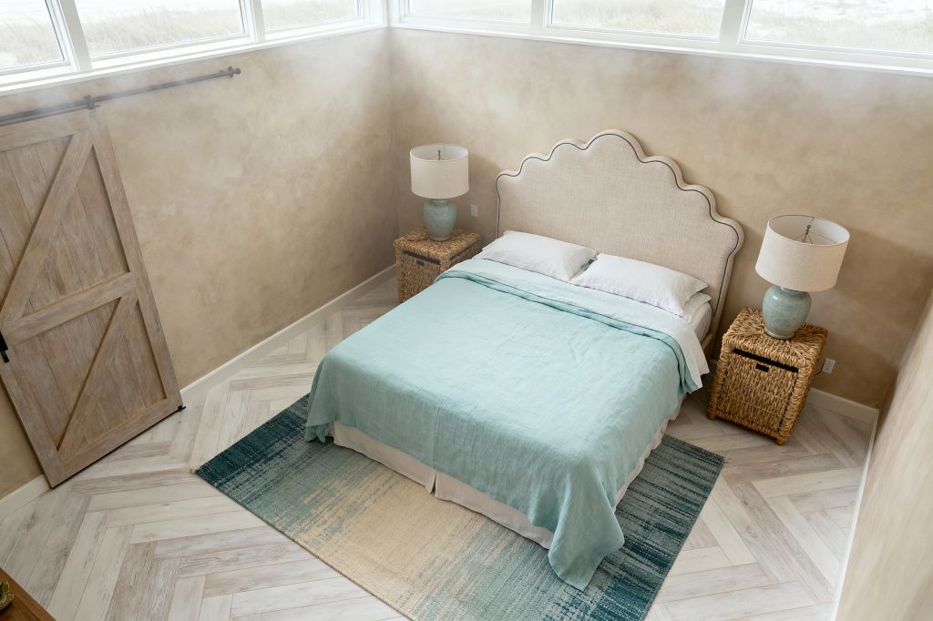 A bright coastal bedroom with a scalloped linen headboard set against sand-colored plaster walls. A queen bed is made with a seafoam linen coverlet over white sheets and a skirt. On each side, woven rattan nightstands hold celadon ceramic lamps with linen drum shades. A teal-to-sand gradient rug anchors the bed on a whitewashed herringbone floor. High clerestory windows wash the room with daylight, and a weathered sliding barn door sits to the left.
