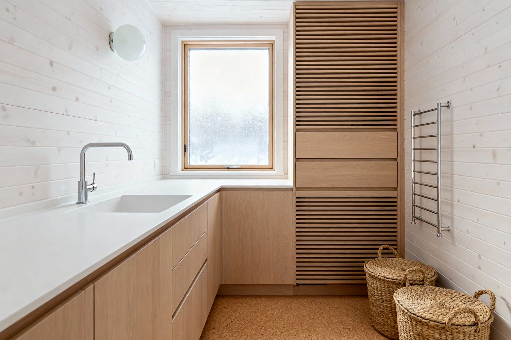 Minimal Scandinavian laundry room with whitewashed wood panel walls and ceiling, a long white countertop with integrated rectangular sink, and flat-front light oak cabinets along the left and back walls. A frosted wood-framed window centers the space, next to a tall slatted oak cabinet, stainless towel rail, and two lidded woven baskets on warm cork flooring, lit by a round opal wall sconce.