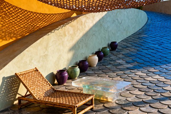 A sunlit patio with a curved stucco wall in ochre and pale sage, where a bronze chain canopy casts netlike shadows across fish-scale pavers. A woven rattan lounge chair sits beside a translucent resin coffee table that glows with refracted light. Along the wall, a neat row of glazed ceramic pots in eggplant, moss green, and cream adds color. The ramping floor and exaggerated shadows create a surreal, dreamlike atmosphere.