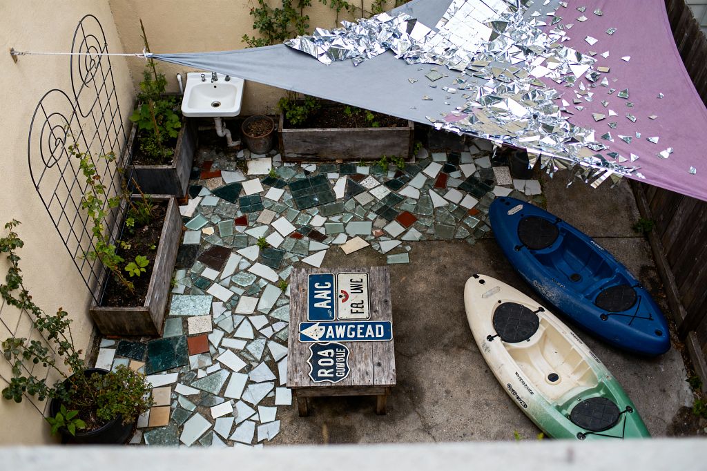 A compact patio viewed from above shows a patchwork floor of broken ceramic and glass tiles in greens, whites, and rust tones beneath a triangular fabric shade sprinkled with reflective mylar pieces. A weathered wood table topped with vintage road signs stands among raised planters, a small wall-mounted sink, and a black metal trellis against beige stucco walls, with blue and white-green sit-on-top kayaks resting on the concrete edge.