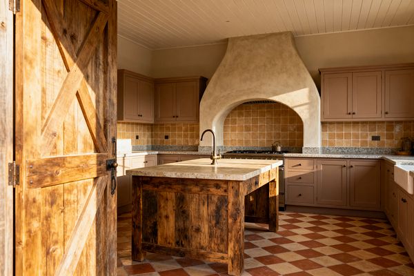 A farmhouse kitchen opens through a barn-style wood door to a U-shaped layout with a central reclaimed-wood island topped in thick stone and a brass gooseneck faucet. Muted clay shaker cabinets run along speckled gray stone counters and a honey-tan square tile backsplash, while a sculpted plaster hood arches over the range. Terracotta-and-cream checkerboard floor tiles and a white tongue-and-groove ceiling glow in warm natural light.